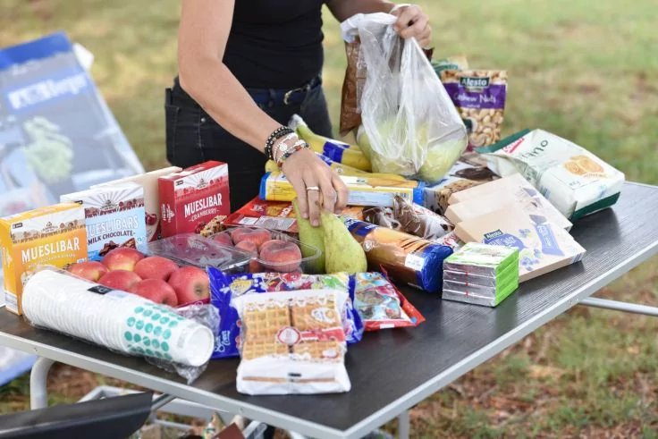 H&auml;nde und Oberk&ouml;rper einer Person legen eine durchsichtige Plastikt&uuml;te mit Birnen auf einen Tisch im Freien. Auf dem Tisch liegen &Auml;pfel, Birnen, M&uuml;sliriegel, Kekspackungen, K&auml;severpackungen, Pappbecher und weitere verpackte Lebensmittel