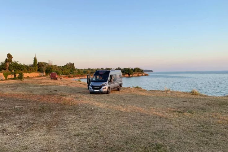 Grau-silberner Campingbus auf trockener Grasfl&auml;che an der K&uuml;ste, ruhiges Meer und klarer Himmel, Person an ge&ouml;ffneter T&uuml;r, B&auml;ume und ein rotes Auto im Hintergrund
