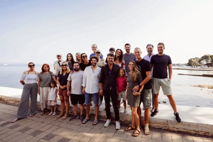 Gro&szlig;e Gruppe von etwa 20 Personen (Erwachsene und Kinder) steht auf einer gepflasterten Promenade vor einer niedrigen Mauer am Meer; heller Himmel im Hintergrund