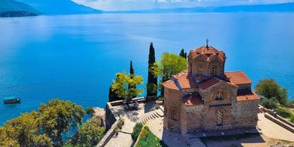 Kleines steinernes Geb&auml;ude mit kuppelf&ouml;rmigem r&ouml;tlichem Ziegeldach auf einer Landzunge am blauen See; B&auml;ume, Treppen und ein kleines Boot im Wasser