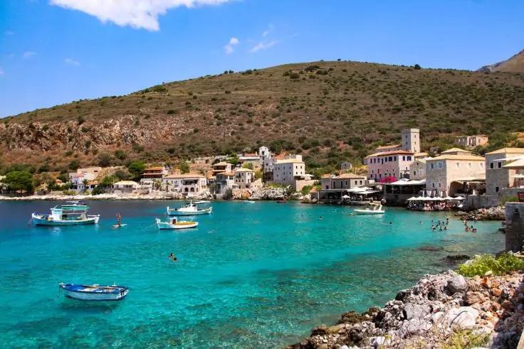 H&auml;userreihe am Ufer einer Bucht mit t&uuml;rkisfarbenem Wasser, mehreren kleinen Booten, felsigem Ufer im Vordergrund und bewachsenem H&uuml;gel im Hintergrund