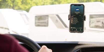 Blick aus dem Fahrersitz auf ein an der Windschutzscheibe befestigtes Smartphone mit sichtbarem Bildschirm; im Vordergrund H&auml;nde am Lenkrad; im Hintergrund geparkte wei&szlig;e Wohnwagen