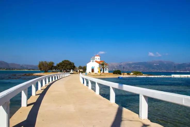 Wei&szlig;e Kapelle mit rotem Dach auf einer kleinen Insel, verbunden durch einen schmalen Fu&szlig;g&auml;ngersteg, blaues Meer und Berge im Hintergrund