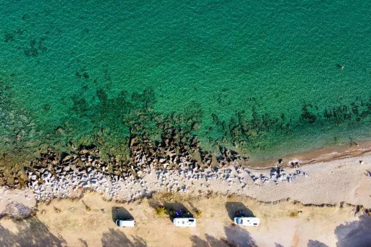Luftaufnahme einer K&uuml;ste mit t&uuml;rkisfarbenem Meer, felsigem Ufer, schmalem Sandstrand und drei wei&szlig;en Wohnmobilen nebeneinander am Strand