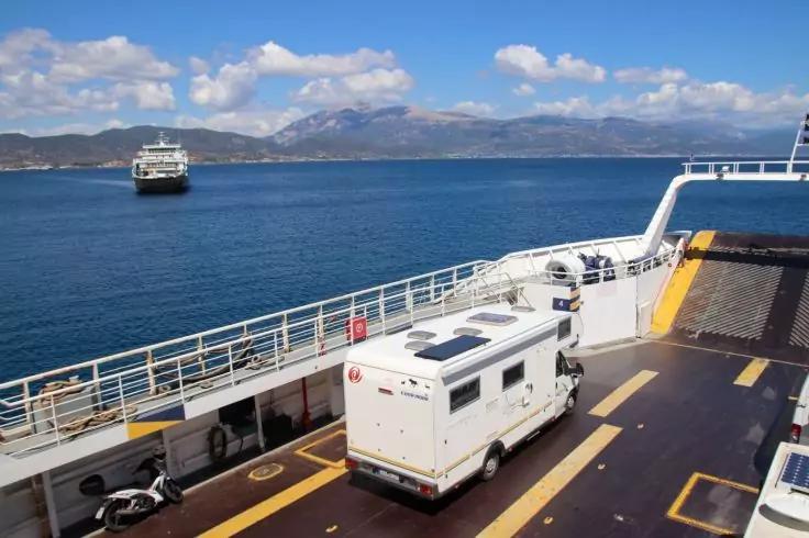 Schiffsdeck mit einem wei&szlig;en Wohnmobil, einem Motorroller, Gel&auml;ndern, einer offenen Fahrzeugrampe, Meer, einem weiteren Schiff in der Ferne und einer bergigen K&uuml;stenlinie unter blauem Himmel mit Wolken