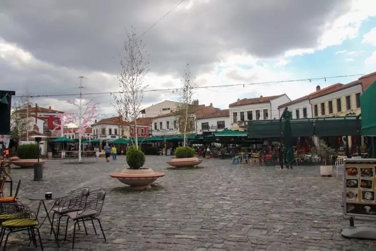 Pflasterplatz mit Caf&eacute;s und Au&szlig;ensitzpl&auml;tzen, Tischen und St&uuml;hlen, gro&szlig;en Pflanzgef&auml;&szlig;en mit B&auml;umen, Menschen, H&auml;userreihe mit roten D&auml;chern und bew&ouml;lktem Himmel
