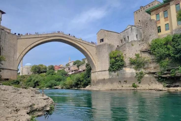 Steinerne Bogenbr&uuml;cke &uuml;ber einen t&uuml;rkisfarbenen Fluss, felsige Uferpartie links, Geb&auml;ude und Steinmauern rechts, B&auml;ume und blauer Himmel