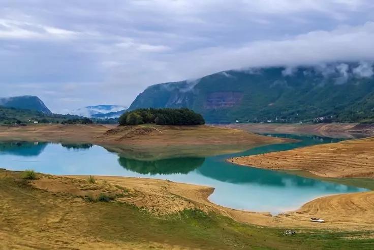 T&uuml;rkisgr&uuml;ne Wasserfl&auml;che mit sandigen Uferpartien, kleine baumbewachsene Insel, gr&uuml;ne Berge im Hintergrund mit niedrigen Wolken, bew&ouml;lkter Himmel