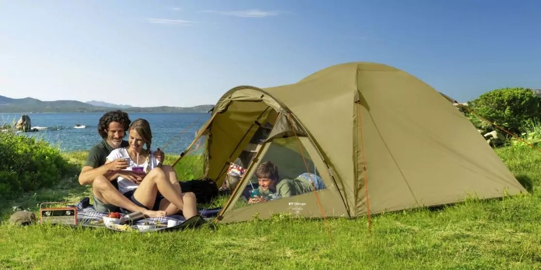 Vier Personen vor einem gr&uuml;nen Zelt auf einer Wiese am Wasser: zwei Erwachsene sitzen auf einer Decke, zwei Kinder liegen im Zelteingang; im Hintergrund Meer, Boote, h&uuml;gelige K&uuml;ste und blauer Himmel