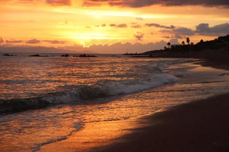 Sonnenuntergang &uuml;ber dem Meer, orangefarbener Himmel mit Wolken, Wellen rollen auf einen sandigen Strand, nasser Sand spiegelt das Licht, Silhouetten von Palmen und Felsen an der fernen K&uuml;ste