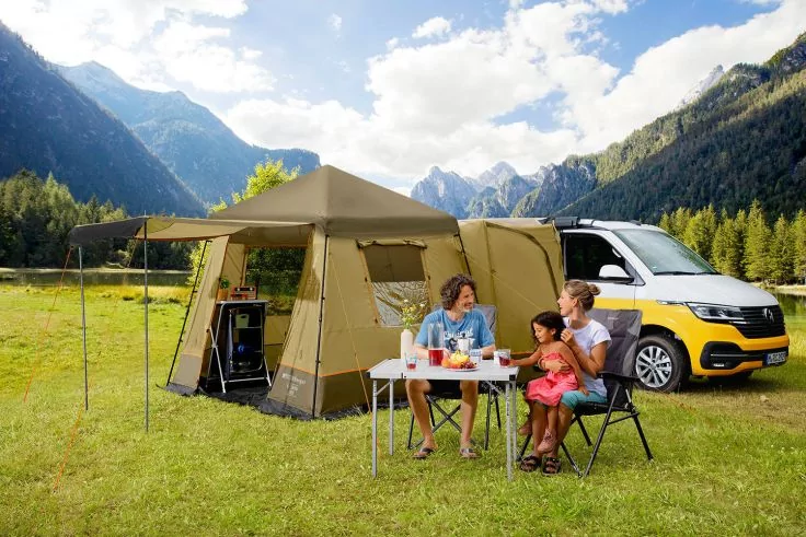 Zeltanbau neben gelb-weißem Campingvan auf einer Wiese; drei Personen sitzen an einem Campingtisch mit Speisen und Getränken, Berge, Bäume und bewölkter Himmel im Hintergrund
