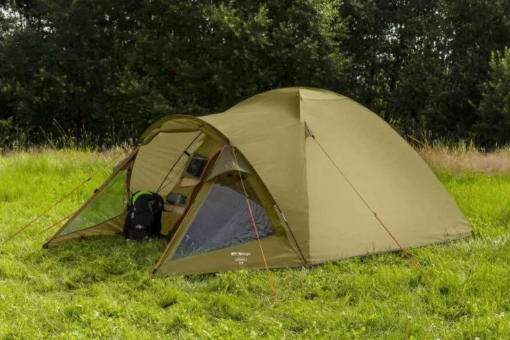 Olivgrünes Kuppelzelt mit offenem Vorraum und einem Rucksack auf Gras vor einer Baumreihe