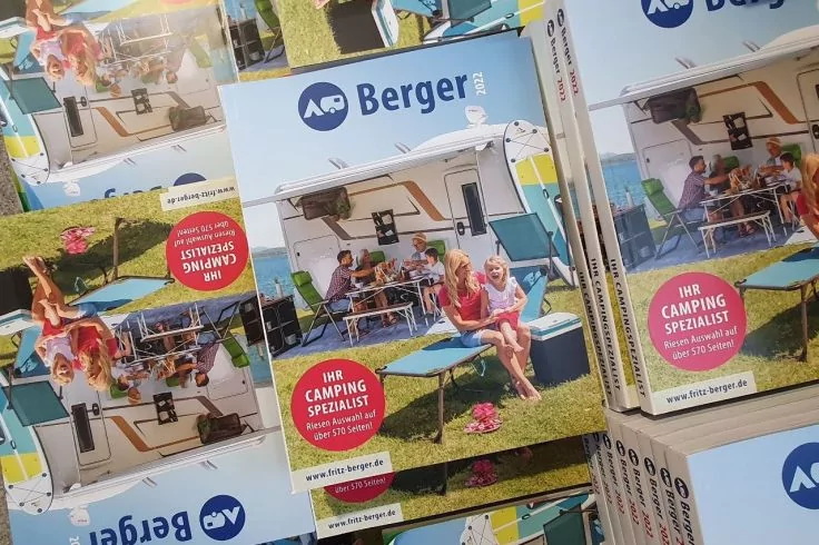 Mehrere gestapelte Broschüren mit dem Titel "Berger" und einem Foto: Wohnwagen, zwei Personen (Erwachsene und Kind) sitzen an einem Tisch auf einer Wiese, roter Aufkleber "IHR CAMPING SPEZIALIST"