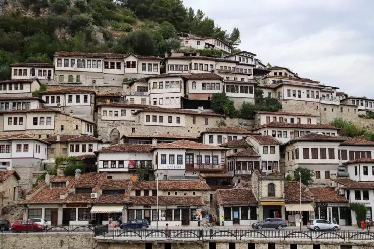 Mehrere weiße Häuser mit Holzfenstern und roten Ziegeldächern dicht an einem Hang mit Bäumen; Straße mit geparkten Autos und Ufermauer im Vordergrund; bewölkter Himmel