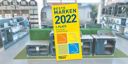 Gelbes Aufsteller-Schild mit dem Text "BESTE MARKEN 2022 1. PLATZ" vor mehreren grauen Kunststoffboxen mit Aussparungen auf gr&uuml;nem Untergrund, im Hintergrund Regale mit Produkten