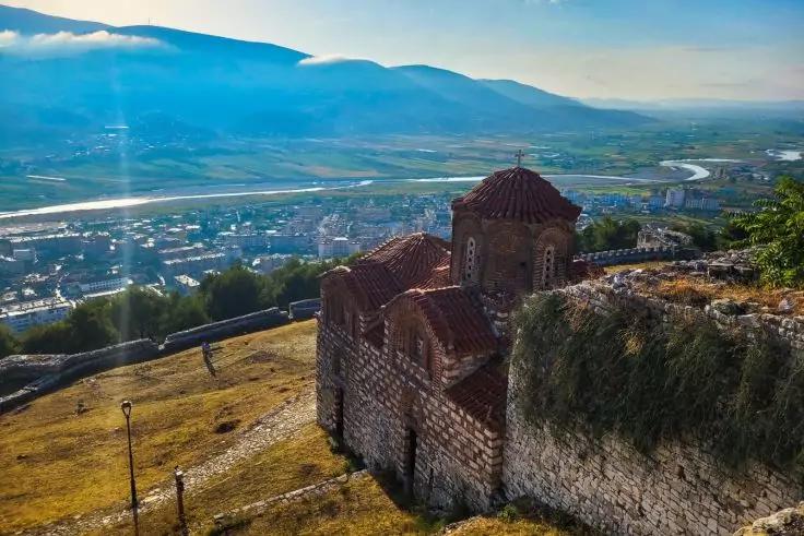 Steinkirche und Mauer auf einem Hügel mit Blick auf Tal, Fluss, Stadt und Berge unter blauem Himmel