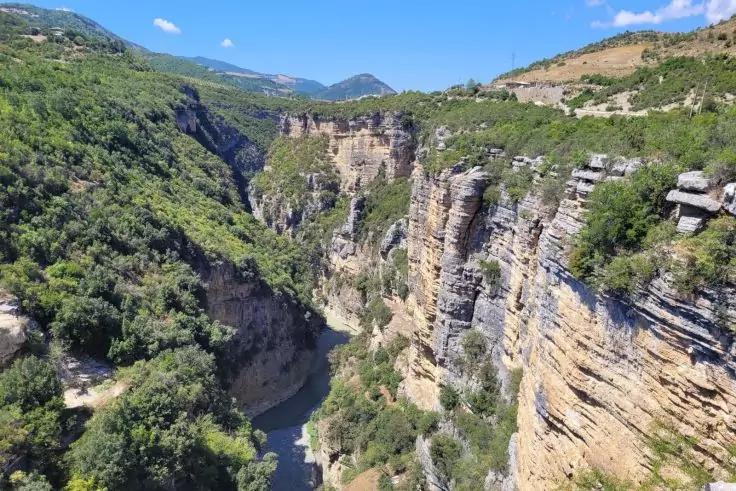 Canyon mit geschichteten Felswänden, Fluss am Boden, grüne Vegetation an den Hängen, Berge im Hintergrund und blauer Himmel mit vereinzelten Wolken