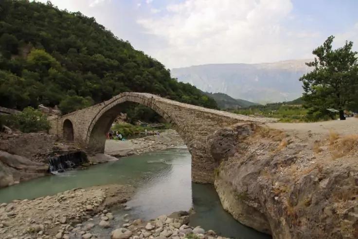 Steinerne einbogige Brücke über einen Fluss, links bewaldeter Hang, rechts felsige Uferzone, im Hintergrund Berge und bewölkter Himmel