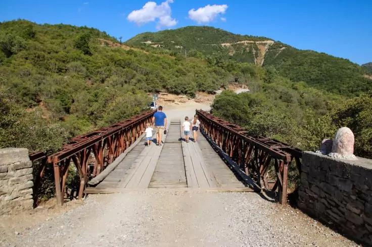 Drei Personen gehen von der Kamera weg über eine Brücke mit Holzplanken und Metallgeländern; links und rechts Steinmauern, dahinter grüne Hügel und blauer Himmel mit einigen Wolken