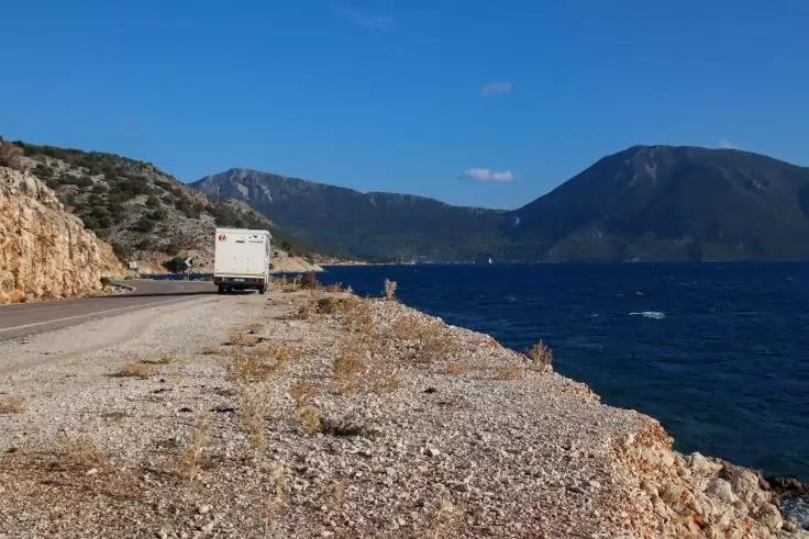 Weißes Wohnmobil an einer Küstenstraße; rechts kiesiger Uferstreifen und Meer; im Hintergrund Berge unter blauem Himmel