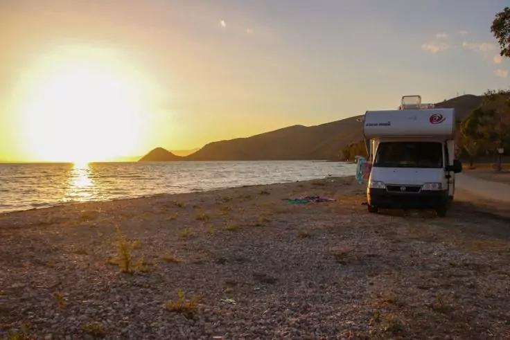 Wohnmobil am kiesigen Strand bei Sonnenuntergang, Meer mit Sonne nahe dem Horizont, Hügel im Hintergrund