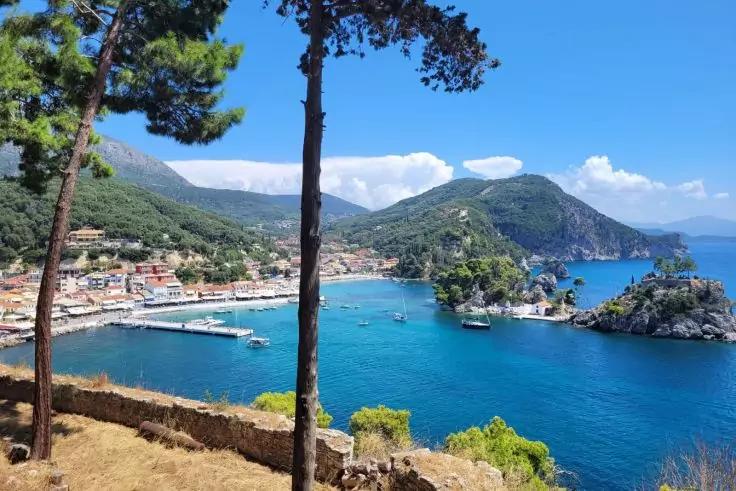 Blick auf eine Küstenbucht mit türkisblauem Wasser, Hafen mit Anlegesteg und Booten, Uferhäuser, felsige Hügel und zwei hohe Kiefern im Vordergrund
