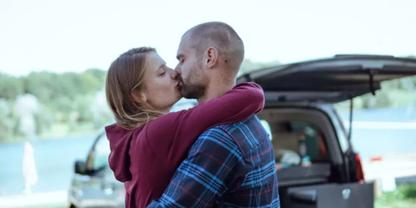 Zwei Personen umarmen sich und k&uuml;ssen sich vor einem ge&ouml;ffneten Kofferraum, im Hintergrund See und B&auml;ume