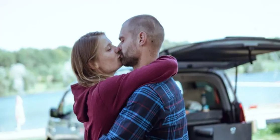 Zwei Personen umarmen sich und k&uuml;ssen sich vor einem ge&ouml;ffneten Kofferraum, im Hintergrund See und B&auml;ume