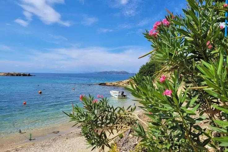 Kieselstrand mit ruhigem türkisfarbenem Meer, kleines weißes Boot nahe der Küste, blühender Oleander mit rosa Blüten im Vordergrund rechts, felsige Landzunge und blauer Himmel im Hintergrund