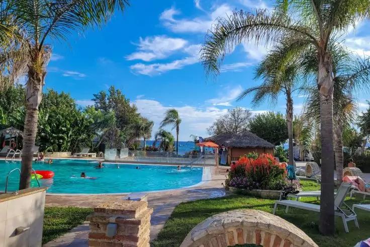 Außen-Schwimmbad mit blauem Wasser, Palmen, Liegestühlen, Personen im Wasser und auf Liegen, Pavillon mit Strohdach, Rasen und steinerner Bogen im Vordergrund, blauer Himmel mit Wolken