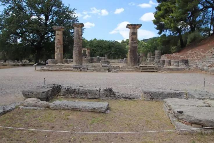 Vier stehende steinerne Säulen auf einem flachen Steinpodest, davor niedrige Steinblöcke und eine Seilabsperrung, im Hintergrund Bäume und blauer Himmel mit wenigen Wolken