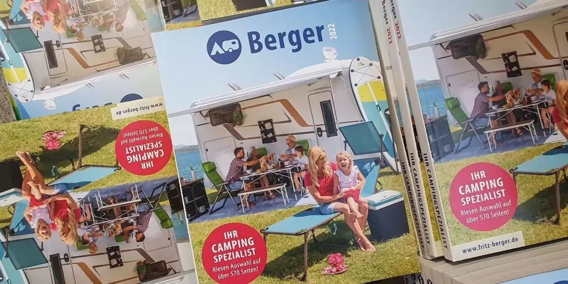 Stapel von Prospekten mit Aufdruck "Berger", Titelbild zeigt wei&szlig;en Wohnwagen mit Markise, zwei Personen sitzen an einem Tisch im Vordergrund, roter runder Aufkleber mit wei&szlig;er Schrift "IHR CAMPING SPEZIALIST"