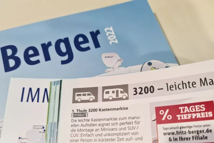 Nahaufnahme mehrerer gedruckter Brosch&uuml;ren auf hellem Untergrund. Vordergrund: wei&szlig;e Seite mit Piktogrammen (Bus, Stra&szlig;enbahn), Text '3200 &ndash; leichte Ma...' und rotem Feld mit Prozentzeichen und 'TAGES TIEFPREISE'. Hintergrund: blaue Brosch&uuml;re mit Sch
