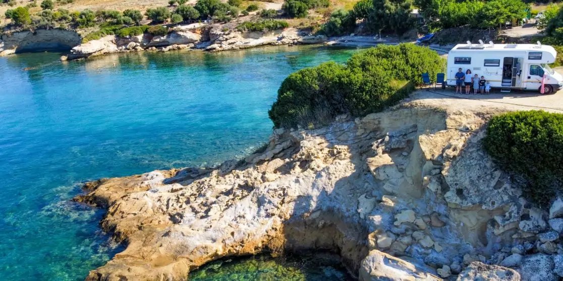 T&uuml;rkisfarbenes Meer neben felsigen Klippen, oben gr&uuml;ne B&uuml;sche und ein wei&szlig;es Geb&auml;ude am Klippenrand