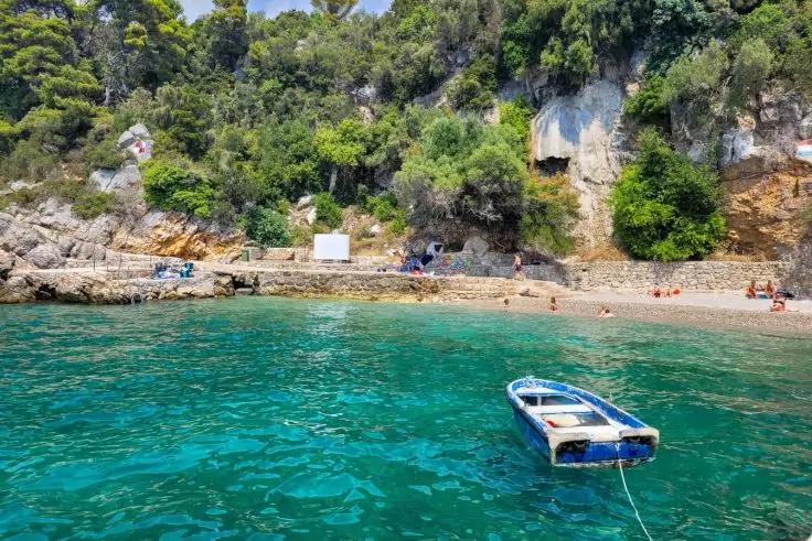 Kleines blaues Boot im Vordergrund auf türkisfarbenem Wasser, dahinter Kiesstrand mit wenigen Menschen und felsige, bewaldete Klippen