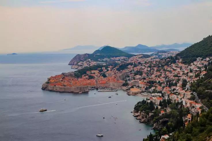 Blick auf eine Küstenstadt mit dicht stehenden Gebäuden und roten Ziegeldächern, einer steinernen Stadtmauer am Ufer, mehreren Booten im Wasser, bewaldeten Hügeln im Hintergrund und hellem Himmel mit leichter Bewölkung