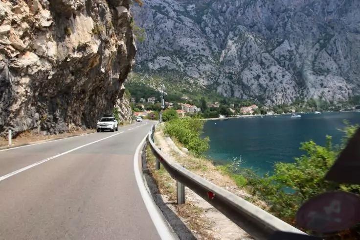 Asphaltierte Küstenstraße mit Leitplanke, weißem Auto links, Felswand links, blaues Wasser und Häuser gegenüber in der Bucht, Berge im Hintergrund