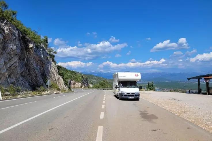 Weißes Wohnmobil am rechten Fahrbahnrand einer asphaltierten Straße; links Felswand, rechts kleiner Parkplatz mit überdachtem Unterstand; blauer Himmel mit weißen Wolken und Bergen am Horizont
