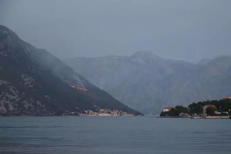 Bucht mit ruhigem Wasser, Berge im Hintergrund, kleines Dorf mit Lichtern am Ufer, Bäume und einige Boote am rechten Ufer