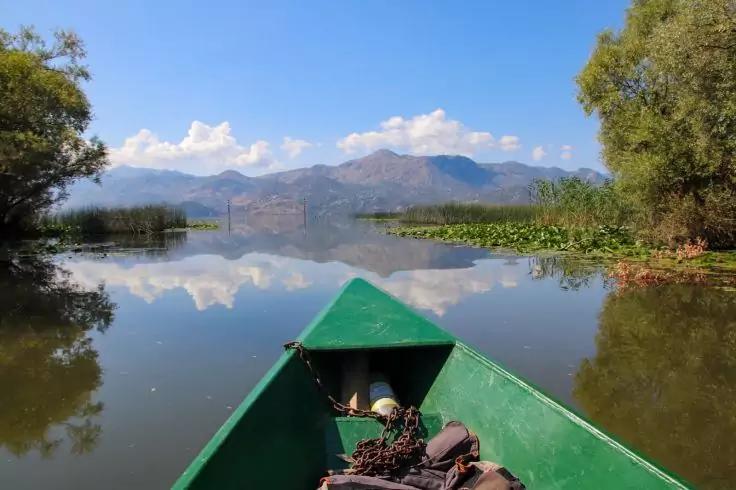 Bug eines grünen Bootes auf ruhigem See mit Spiegelung von Wolken und Bergen, Ufervegetation links und rechts