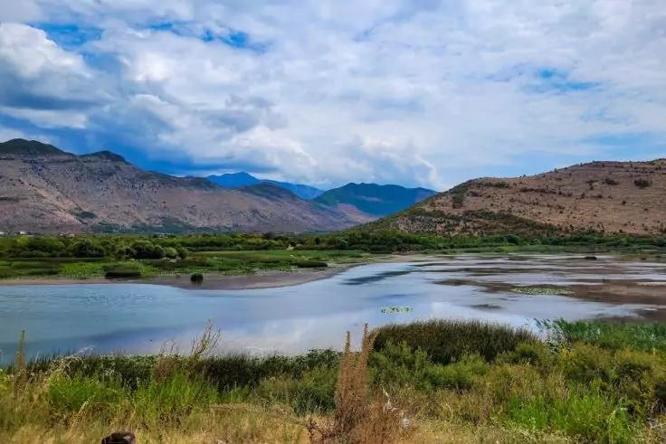 Flaches Gewässer mit Schilf und Gras im Vordergrund, Hügel und bewölkter Himmel im Hintergrund