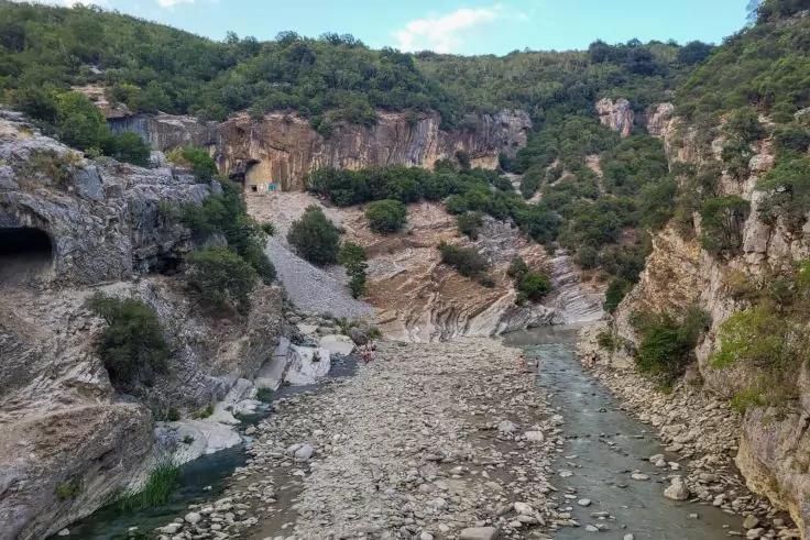 Fluss mit schmalem Wasserlauf und steinigem Flussbett, umgeben von steilen Felswänden, vereinzelten Büschen und Bäumen, blauer Himmel mit wenigen Wolken