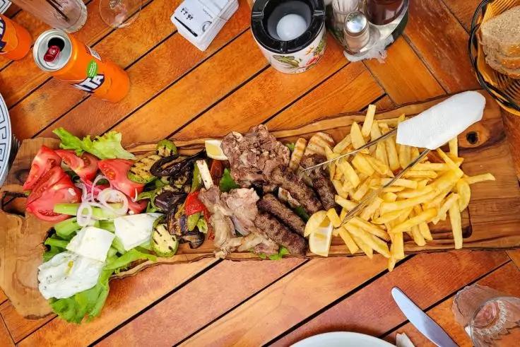 Holzbrett mit Fleischstücken, Pommes Frites, grünen Salatblättern, Tomatenscheiben, Zwiebelringen, weißen Käsewürfeln, einer Zitronenscheibe, einer Serviette, Gewürzstreuern und Besteck auf einem Holztisch