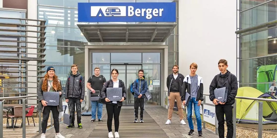 Acht Personen stehen auf einem Holzsteg vor dem gl&auml;sernen Eingang eines Geb&auml;udes mit blauem "Berger"-Schild; mehrere halten Laptops oder Ordner; links Tische und St&uuml;hle, rechts ein gr&uuml;nes Zelt