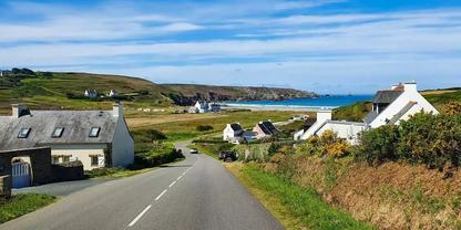 Landstra&szlig;e f&uuml;hrt bergab zu einer Sandbucht; wei&szlig;e H&auml;user links und rechts, gr&uuml;ne Wiesen, Felsklippen am Meer, blaues Meer und wolkiger Himmel