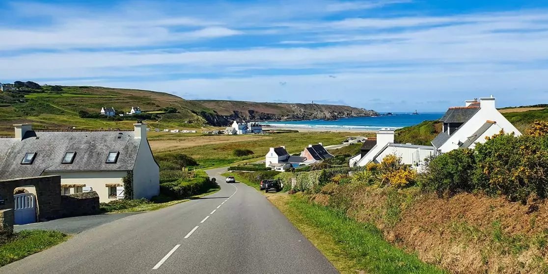 Landstra&szlig;e f&uuml;hrt bergab zu einer Sandbucht; wei&szlig;e H&auml;user links und rechts, gr&uuml;ne Wiesen, Felsklippen am Meer, blaues Meer und wolkiger Himmel