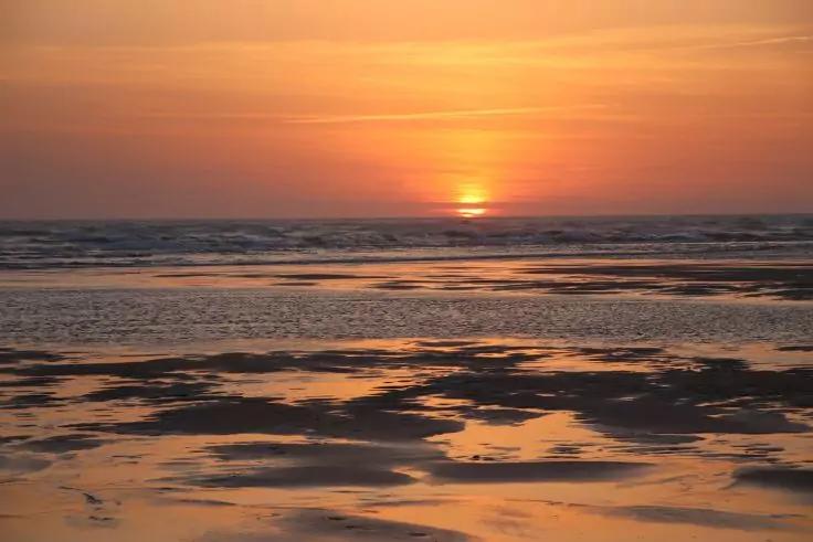 Sonne knapp &uuml;ber dem Horizont &uuml;ber dem Meer, oranger Himmel und spiegelnde Pf&uuml;tzen auf nassem Sand bei Ebbe