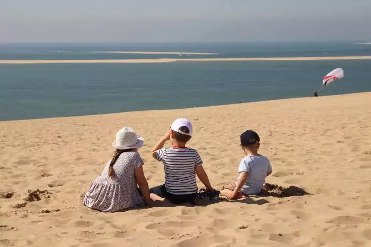 Drei Kinder sitzen auf einer Sandd&uuml;ne und blicken aufs Meer; links ein Kind mit Sonnenhut, in der Mitte ein Kind mit wei&szlig;er Kappe und gestreiftem T-Shirt, rechts ein Kind mit dunkler Kappe. Im Hintergrund &uuml;ber dem Wasser ein rot-wei&szlig;es Fallschirmsege