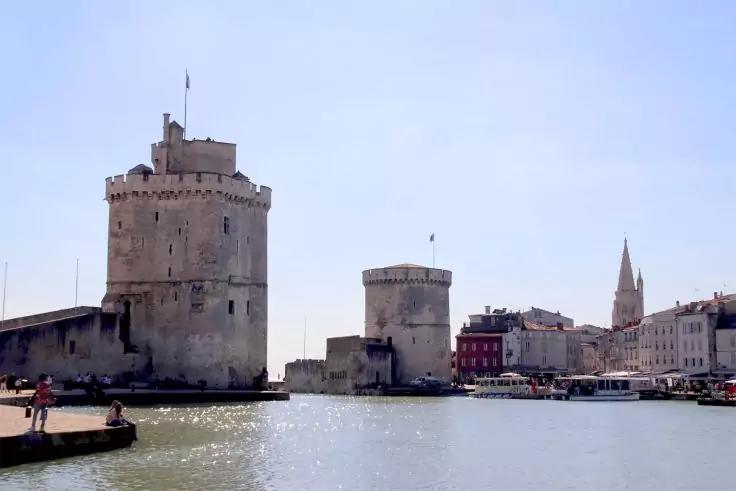 Zwei runde Steint&uuml;rme an einem Hafen, Wasser im Vordergrund, Boote am Kai, Geb&auml;ude und ein Kirchturm im Hintergrund, klarer Himmel