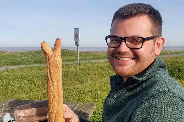 Person mit Brille und gr&uuml;nem Pullover, l&auml;chelt und h&auml;lt zwei lange Baguettes an einem Holztisch; im Hintergrund Gras, ein Schild, Wasser und klarer Himmel